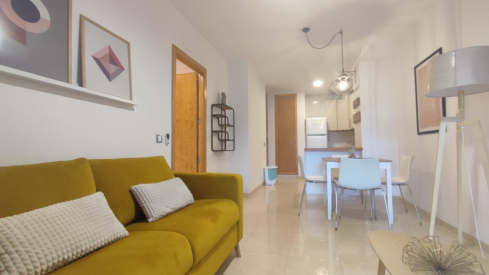 Un salon moderne avec un canapé de couleur moutarde, un coin repas avec des chaises blanches et une cuisine en arrière-plan.