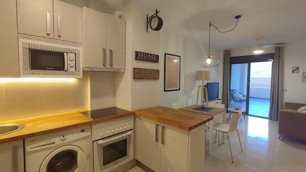 A compact kitchen with a microwave, oven, and washing machine, a wooden countertop, and a dining area with a table and chairs. - La Envía - Beautiful apartment with terrace, swimming pool, luminous (La Envía)