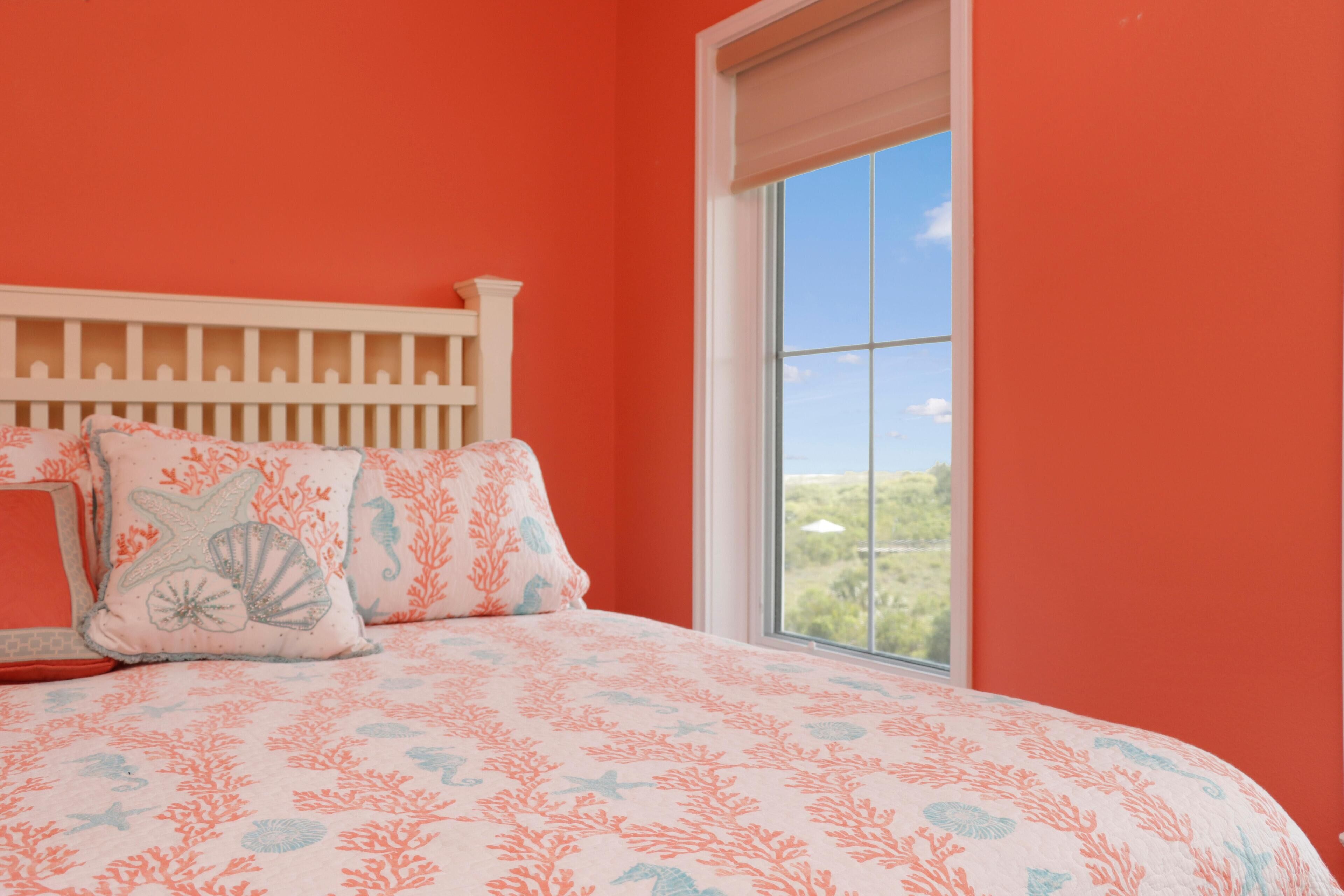 A bedroom with a white headboard, a large window with a view of greenery, and a bed with a coral-patterned bedspread.