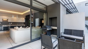 Une cuisine moderne avec une porte vitrée donnant sur un coin repas, un barbecue et un espace détente comprenant un canapé et des fauteuils.