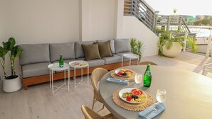 Un espacio al aire libre moderno con un sofá gris, dos mesas redondas pequeñas, sillas de mimbre y plantas en macetas.