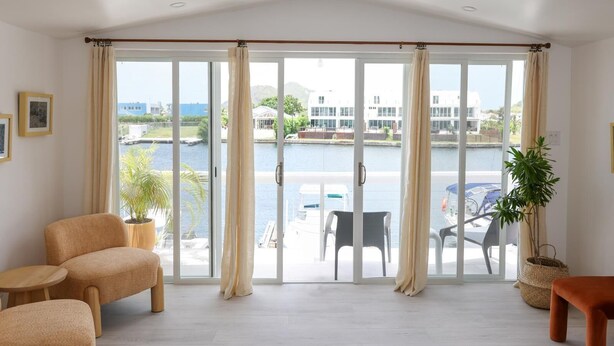 Amplia habitación con un gran ventanal corredero que da a un balcón con vistas a un puerto deportivo y edificios. En la habitación hay dos sillas y una mesita.