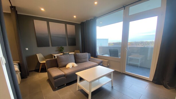 Uma sala de estar moderna com um sofá cinzento, uma mesa de centro branca e uma grande janela com vista para a cidade.