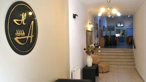 Een hotel lobby met een receptiebalie, een zwarte bank, een witte vaas met bloemen, een trap naar een eetgedeelte en een televisie aan de muur.