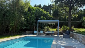 Piscina con sillones de descanso y zona de estar cubierta, rodeada de follaje.