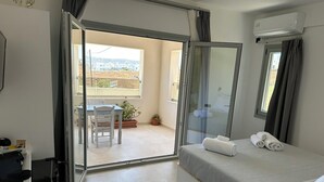 A hotel room with a bed, a desk, a chair, and a balcony with a view. - Lovely Hot-L (Santorini)