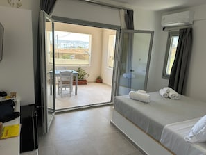A hotel room with a bed, a desk, a chair, and a balcony with a view. - Lovely Hot-L (Santorini)