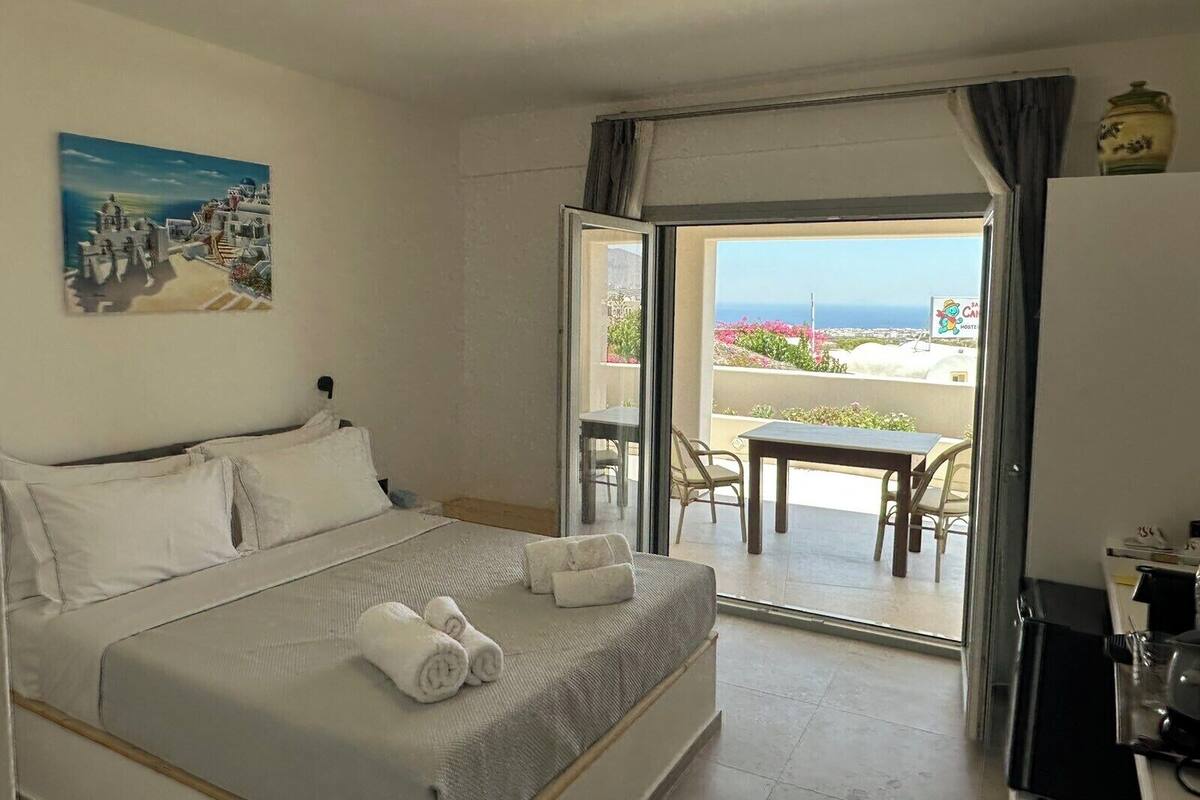 A hotel room with a bed, a painting of a coastal scene, a balcony with a table and chairs, and a view of the sea.