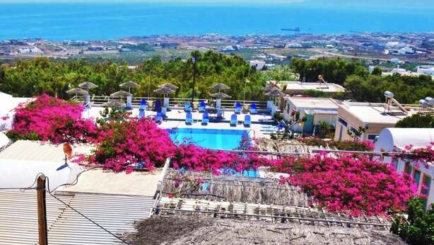 A pool area with lounge chairs and umbrellas, surrounded by pink bougainvillea flowers, with a view of the sea and distant buildings. - Lovely Hot-L (Santorini)