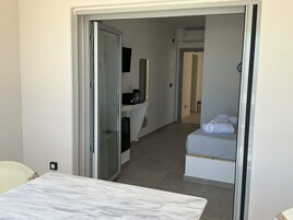 Habitación de hotel con una mesa de mármol, una cama con toallas dobladas, una pequeña kitchenette y un baño a la vista a través de una puerta abierta.