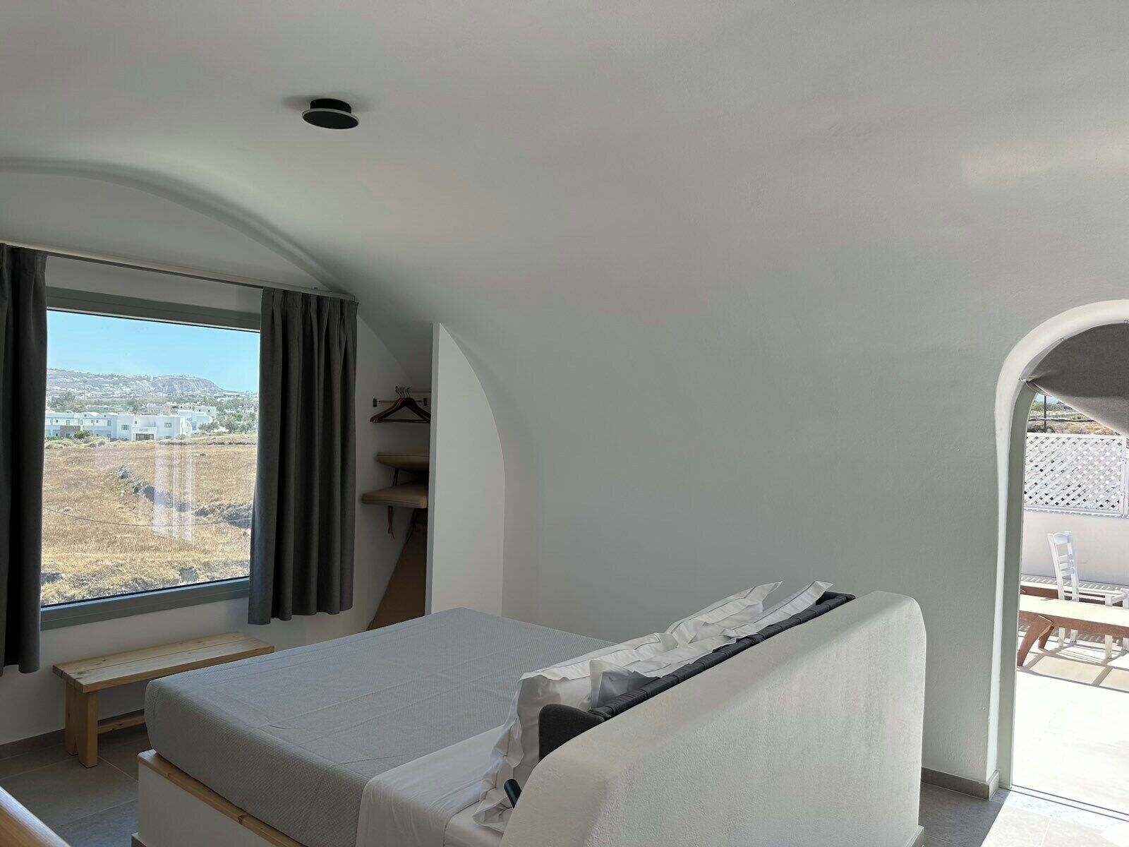 A bedroom with a bed, a bench, a window with curtains, and a view of buildings outside.