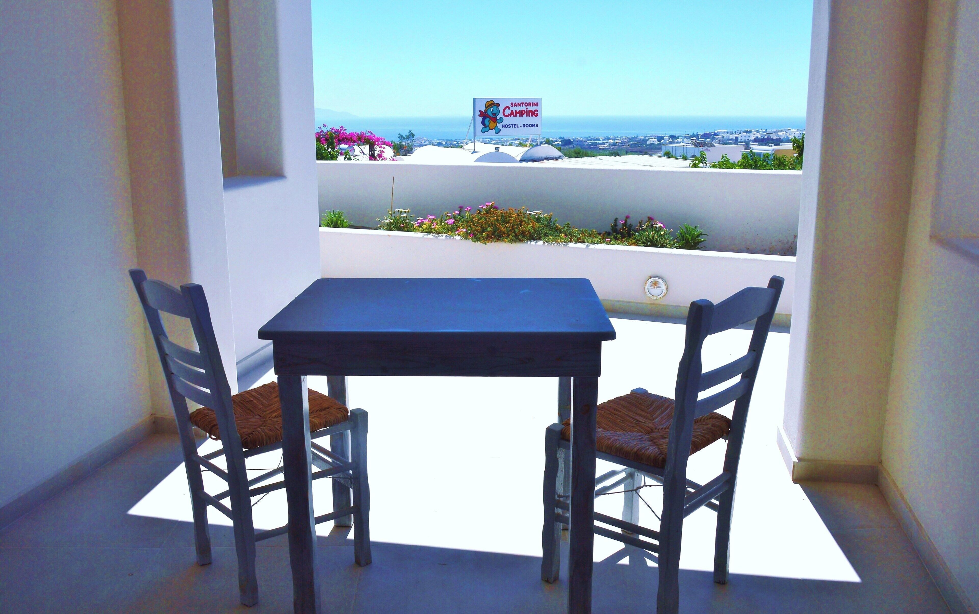 A balcony with a table and two chairs, a view of the sea, and a sign for Santorini Camping Hostel-Rooms.