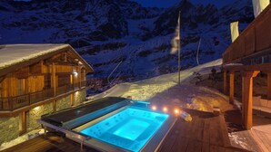 Een houten hut met een jacuzzi op een houten terras, omgeven door met sneeuw bedekte bergen.