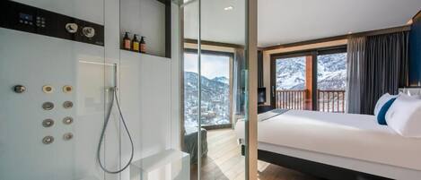 Una habitación de hotel moderna con una cama grande, zona de ducha con mampara de vidrio y un balcón con vistas a la montaña.