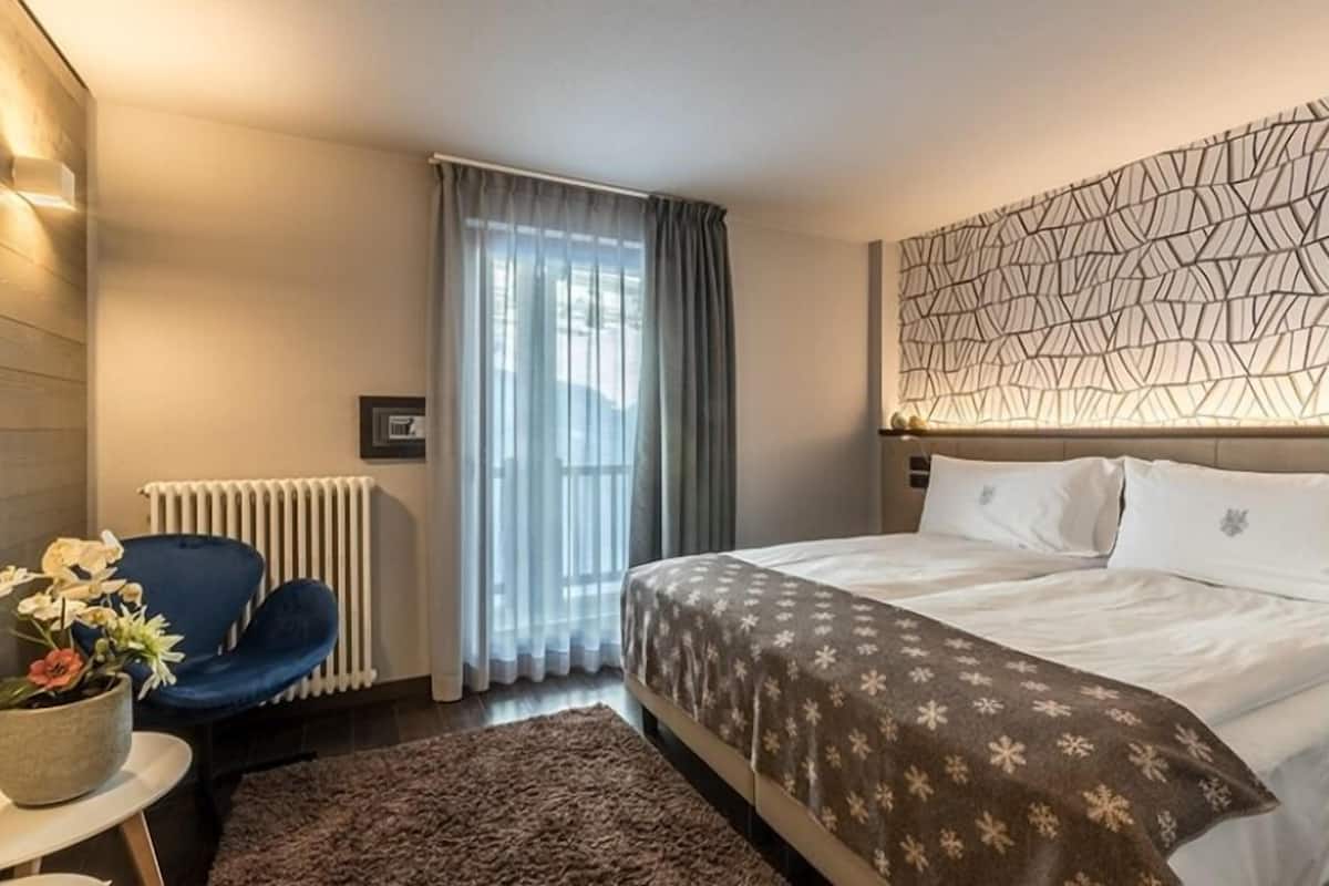 A hotel room with a bed, a chair, a radiator, a window with curtains, and a wall with a geometric pattern.