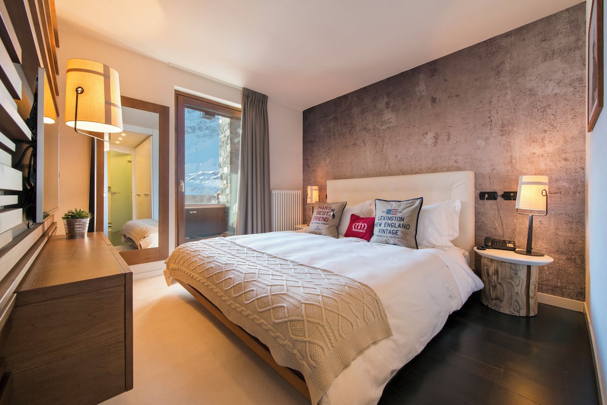 A modern hotel room with a large bed, bedside lamps, a wooden nightstand, and a view of the outdoors.