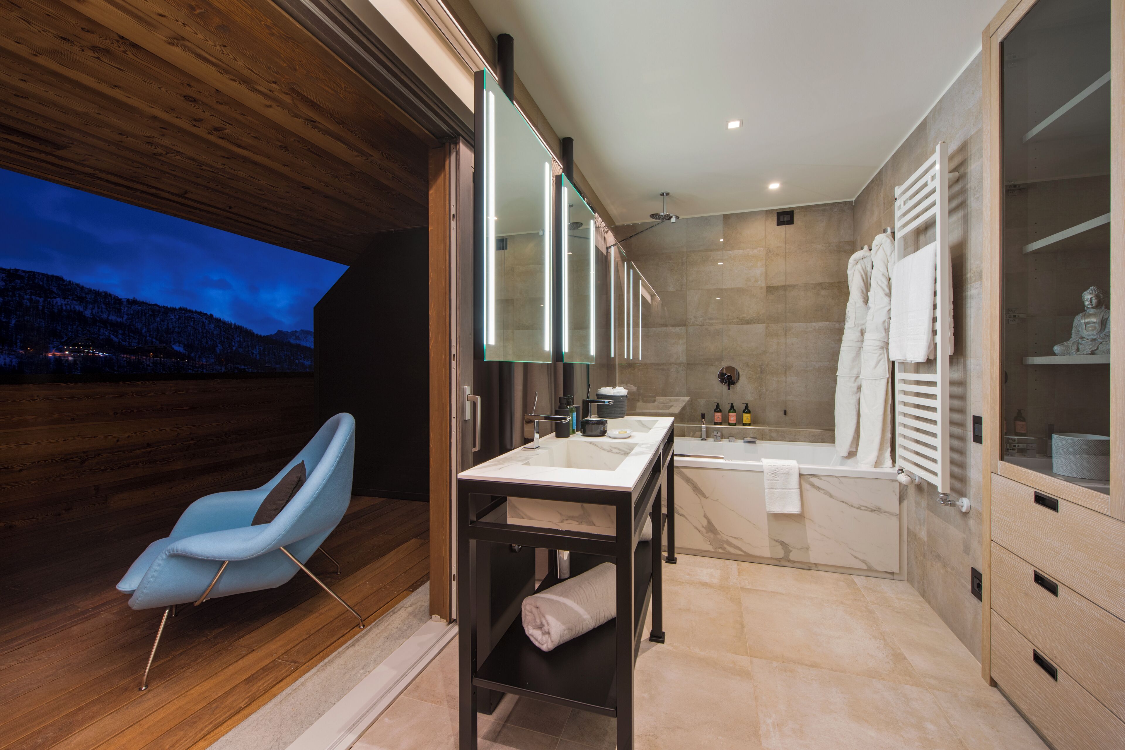 Ein modernes Badezimmer mit Holzboden, einem blauen Stuhl, einem Glastischchen, einer Badewanne und einem großen Spiegel.