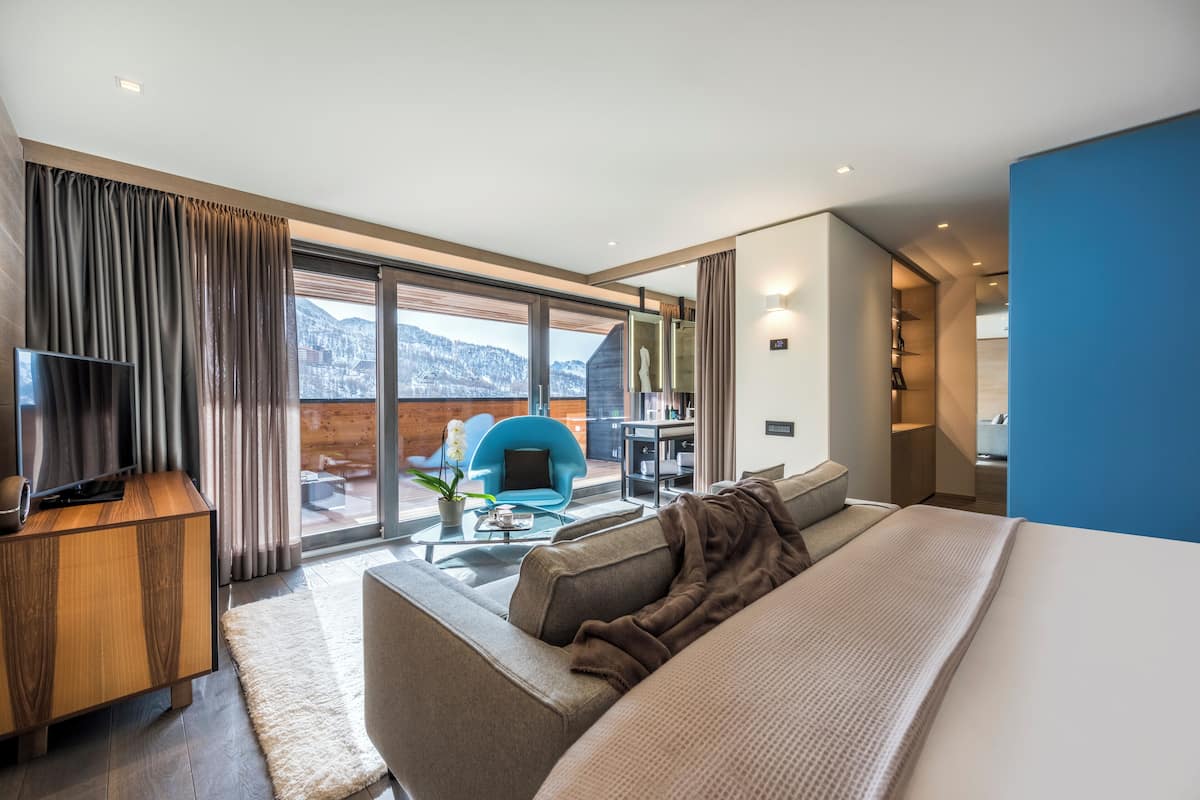 A modern hotel room with a large bed, a TV, a sofa, and a dining area with a view of the mountains.
