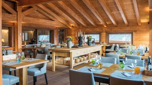 Een houten interieur met hoge plafonds en een centrale eethoek. Er staan tafels klaar met stoelen, en ramen zorgen voor natuurlijk licht.