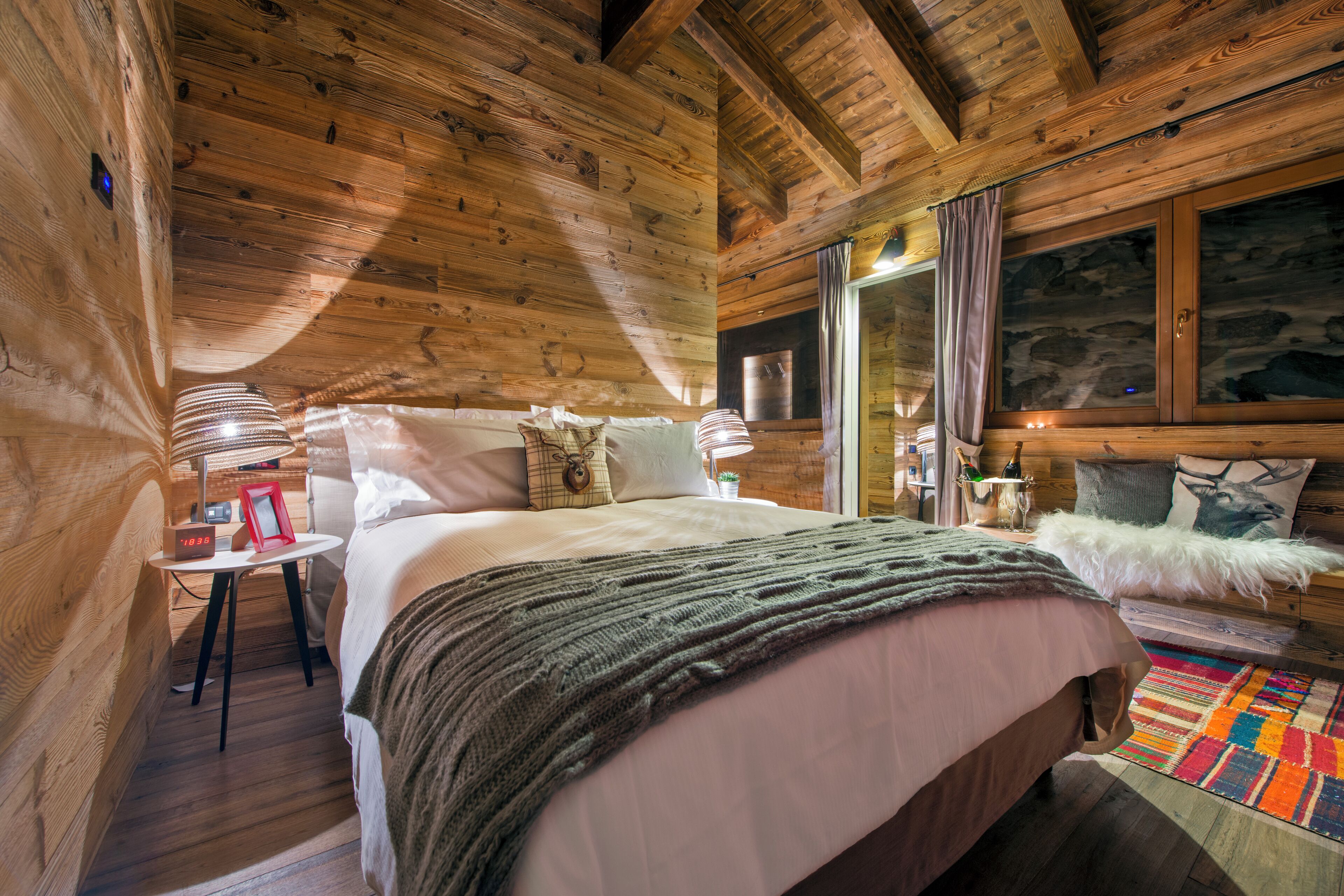 A cozy wooden cabin bedroom with a large bed, a small table with a lamp, and a window with curtains.