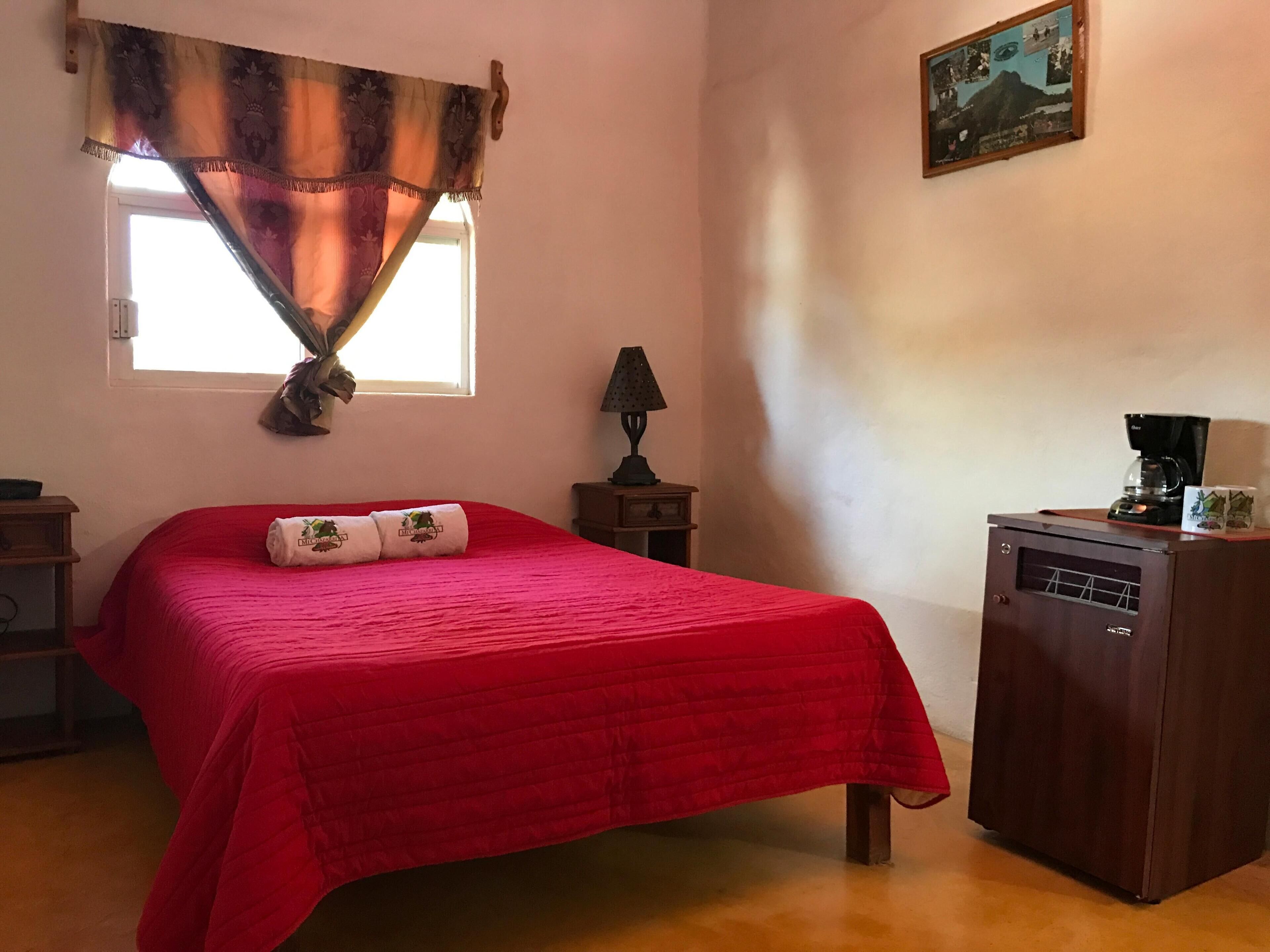 A bedroom with a red bedspread, a small bedside table with a lamp, a coffee maker, and a framed picture on the wall.