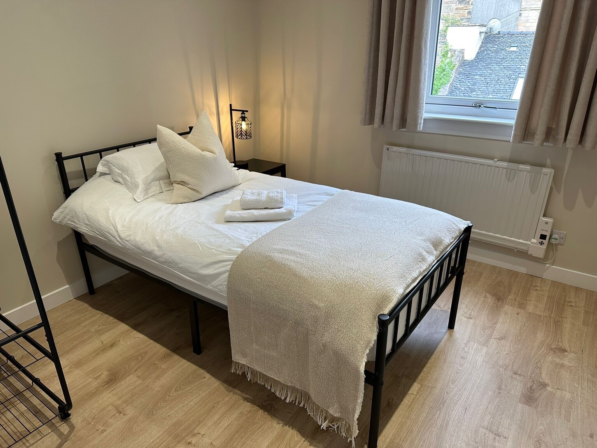 A neatly made bed with white linens and a beige throw blanket, a bedside table with a lamp, and a window with curtains.