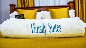A neatly folded white towel with "Vimally Suites" embroidered in blue, placed on a bed with yellow bedding and white pillows. - Vimally Suites Naivasha (Naivasha)