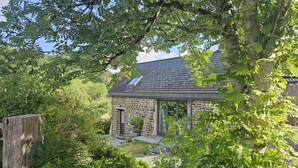 A stone building with a sloped roof and a skylight, surrounded by lush greenery and a wooden fence. - The Stone Barn (Login)