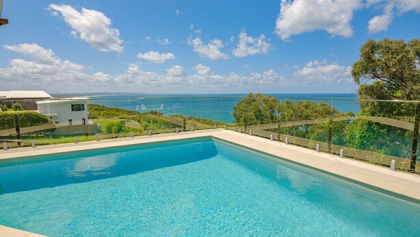 Une piscine à l’eau d’un bleu cristallin, entourée d’une balustrade en verre, offrant une vue panoramique sur l’océan et un ciel dégagé.