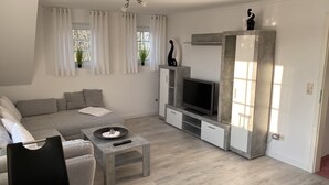 A modern living room with a grey sofa, a coffee table, a TV on a built-in shelf, and a window with curtains. - Apartment 'Adlerhorst' with Shared Terrace, Wi-Fi and Air Conditioning (Wangerland)