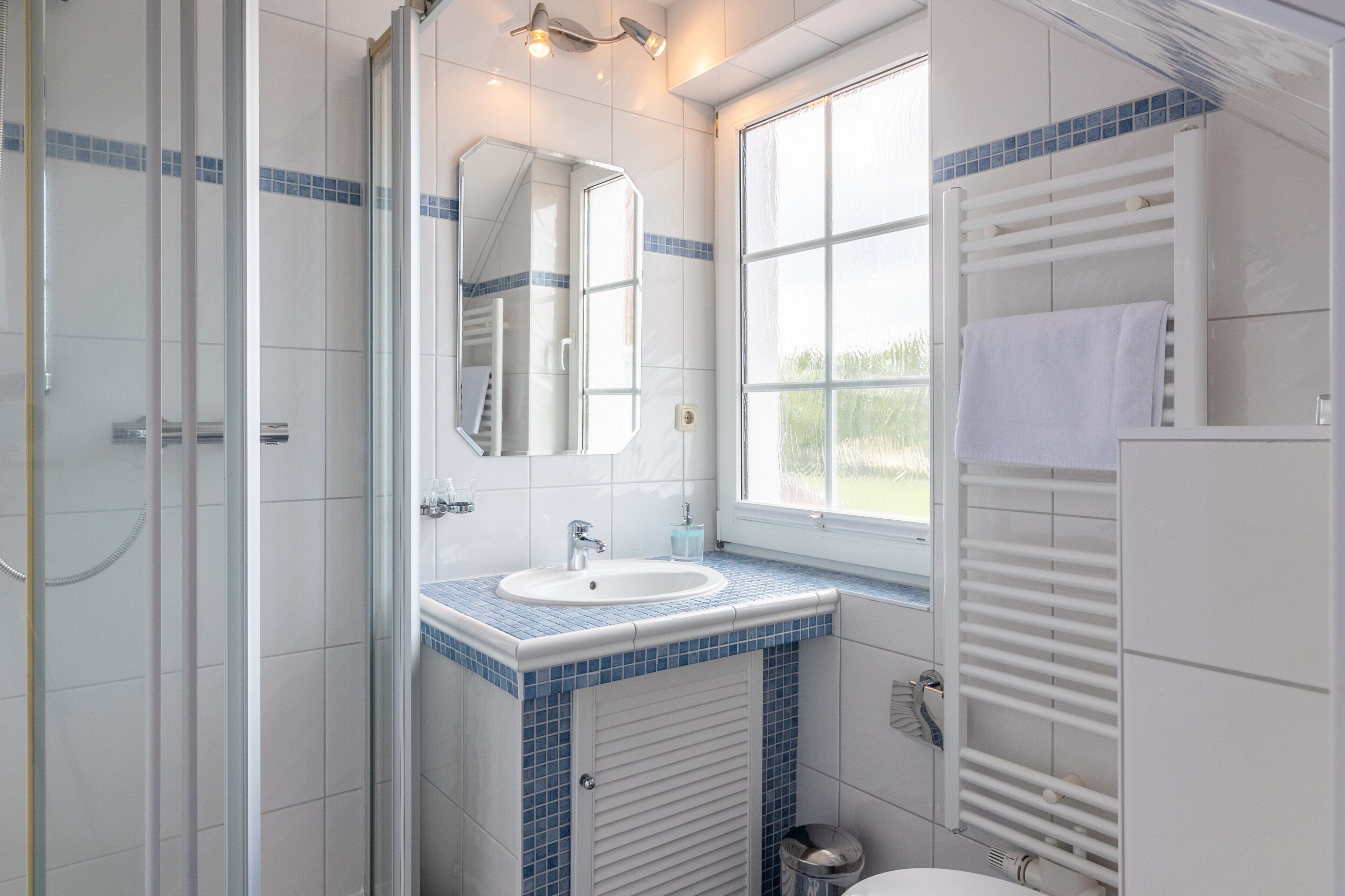 A bathroom with a glass shower enclosure, a sink with blue-tiled countertop, a mirror, and a window.