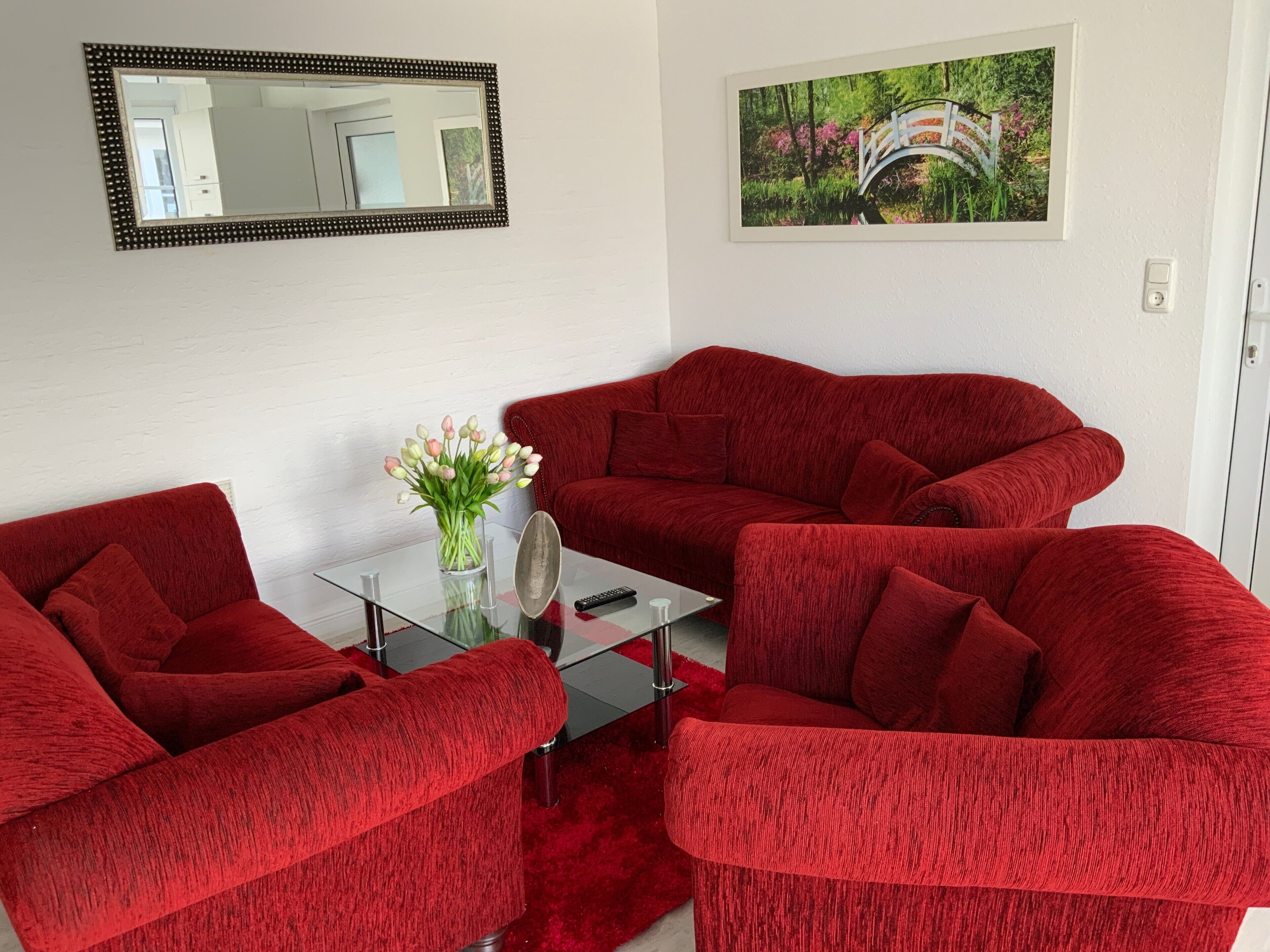 Ein Zimmer mit einer roten Sofagarnitur, einem Glastisch, einer Vase mit Blumen, einem gerahmten Bild an der Wand und einem Spiegel.
