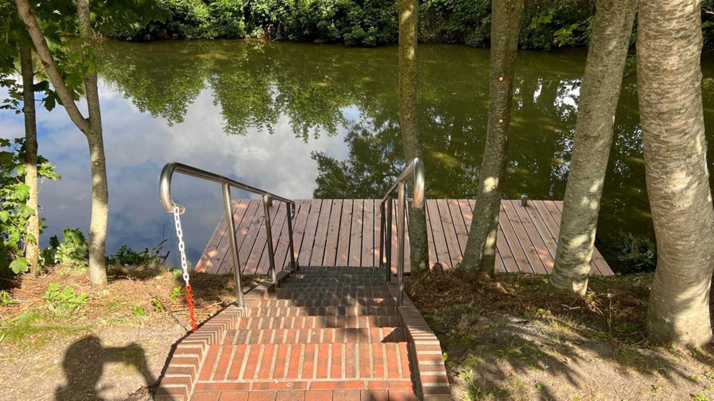 Eine Holzterrasse mit Metallgeländern, die zu einem Gewässer führt, umgeben von Bäumen.