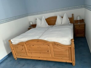 A wooden bed with white bedding, a nightstand with a lamp, and a wooden dresser in a room with blue flooring and white walls.