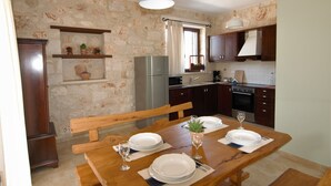A dining area with a wooden table set for four, a refrigerator, and a stone wall. - Rosemary Cottage, Lioriza Houses Zakynthos (Zakynthos)
