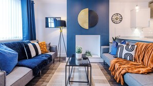A modern living room with a blue accent wall, a sofa, a coffee table, and a side table with a plant. - 3BR House w/ Parking, Free Wifi, and Discounts! (Droylsden)