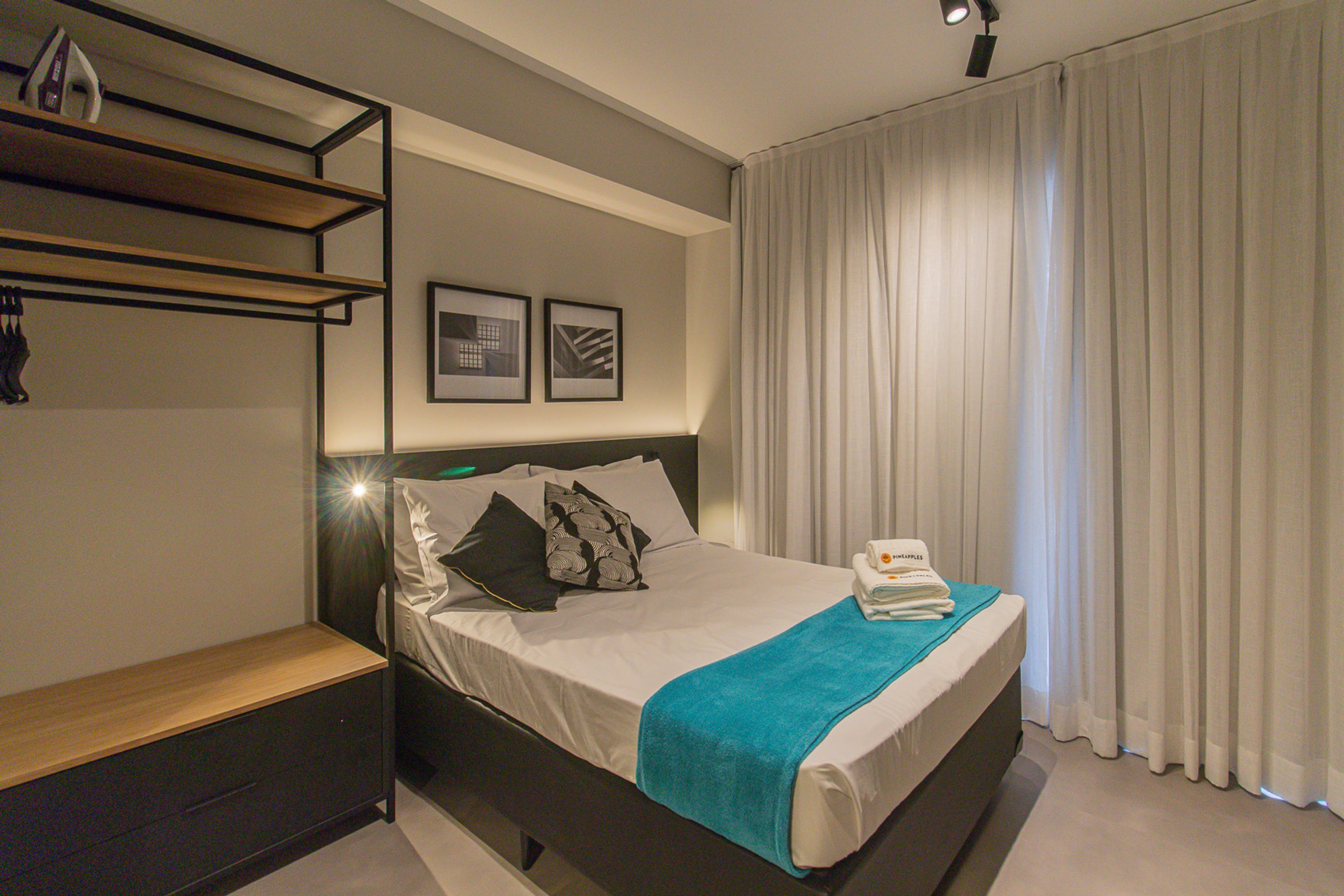 A modern hotel room with a bed, a bedside table, a shelf with items, and a window with curtains.