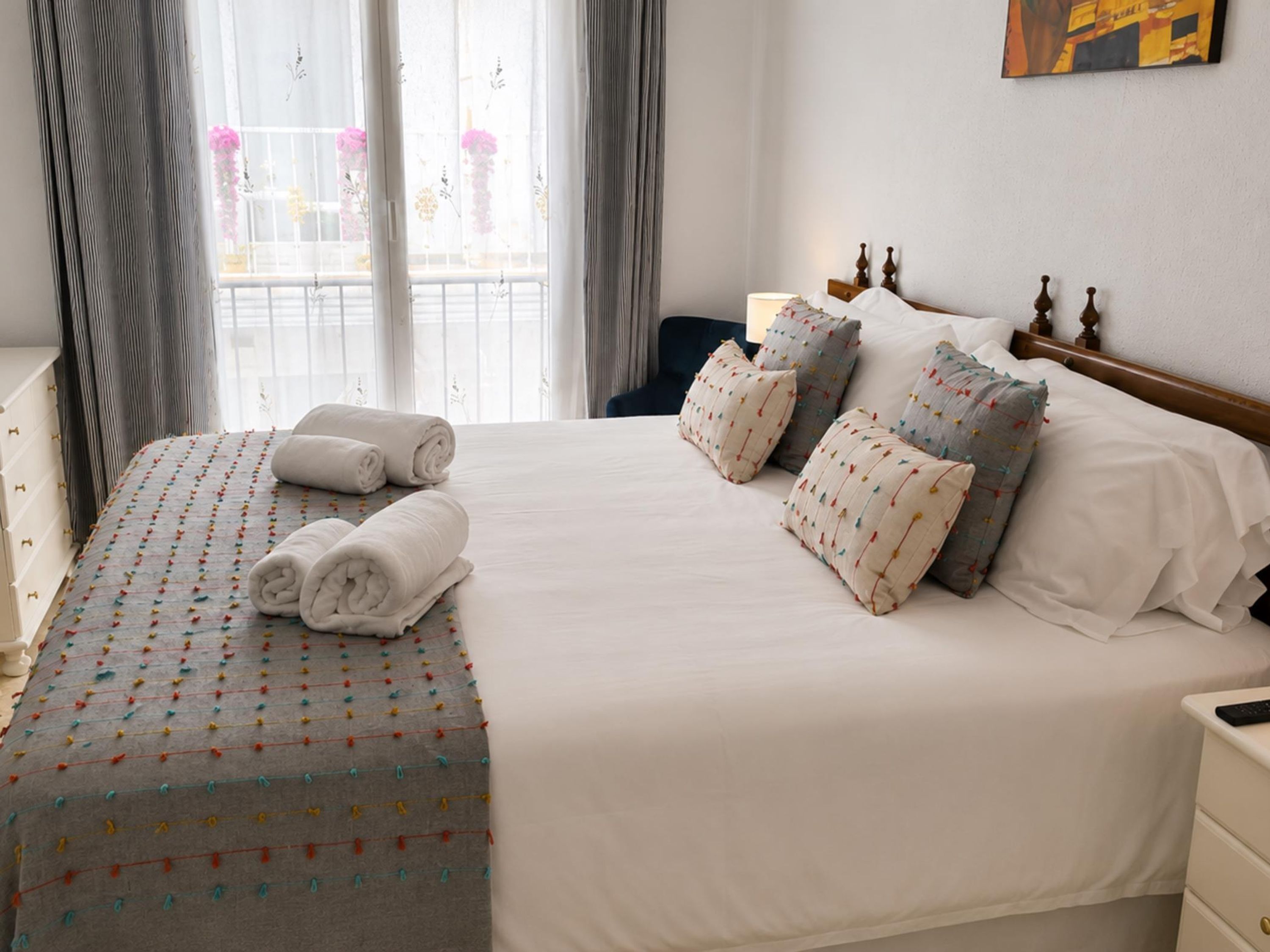 A neatly made bed with pillows and rolled towels, a bedside table with a lamp, and a window with curtains.