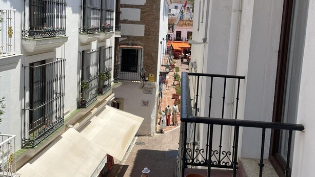 A narrow street with buildings on either side, featuring balconies with metal railings and a small shop at the bottom. - Plaza de los Naranjos (Marbella)
