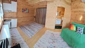 Una habitación tipo cabaña de madera con una cama verde, una kitchenette pequeña, un baño y un cuadro en la pared.