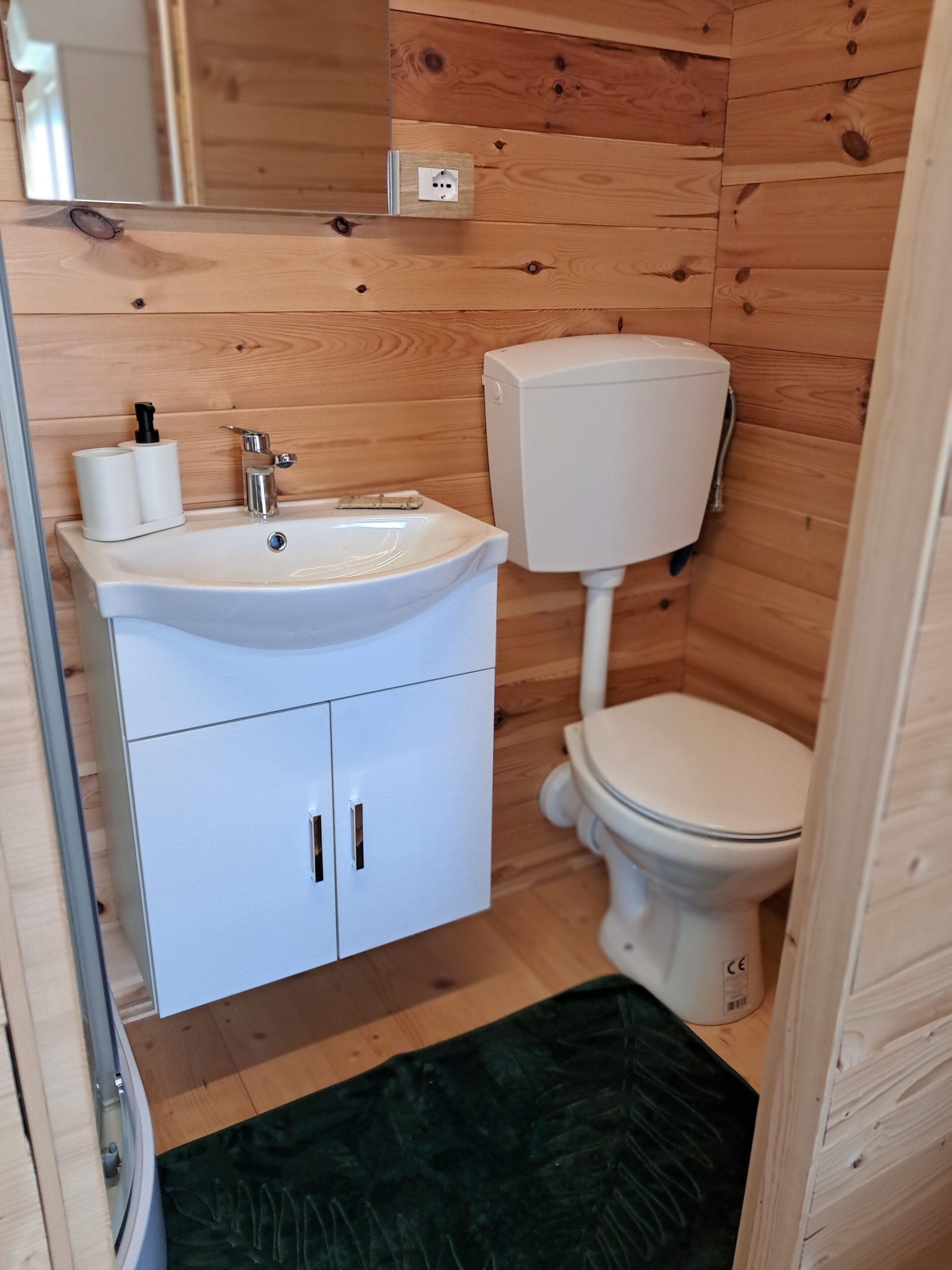A small bathroom with a wooden wall, a white sink, a toilet, and a green rug.