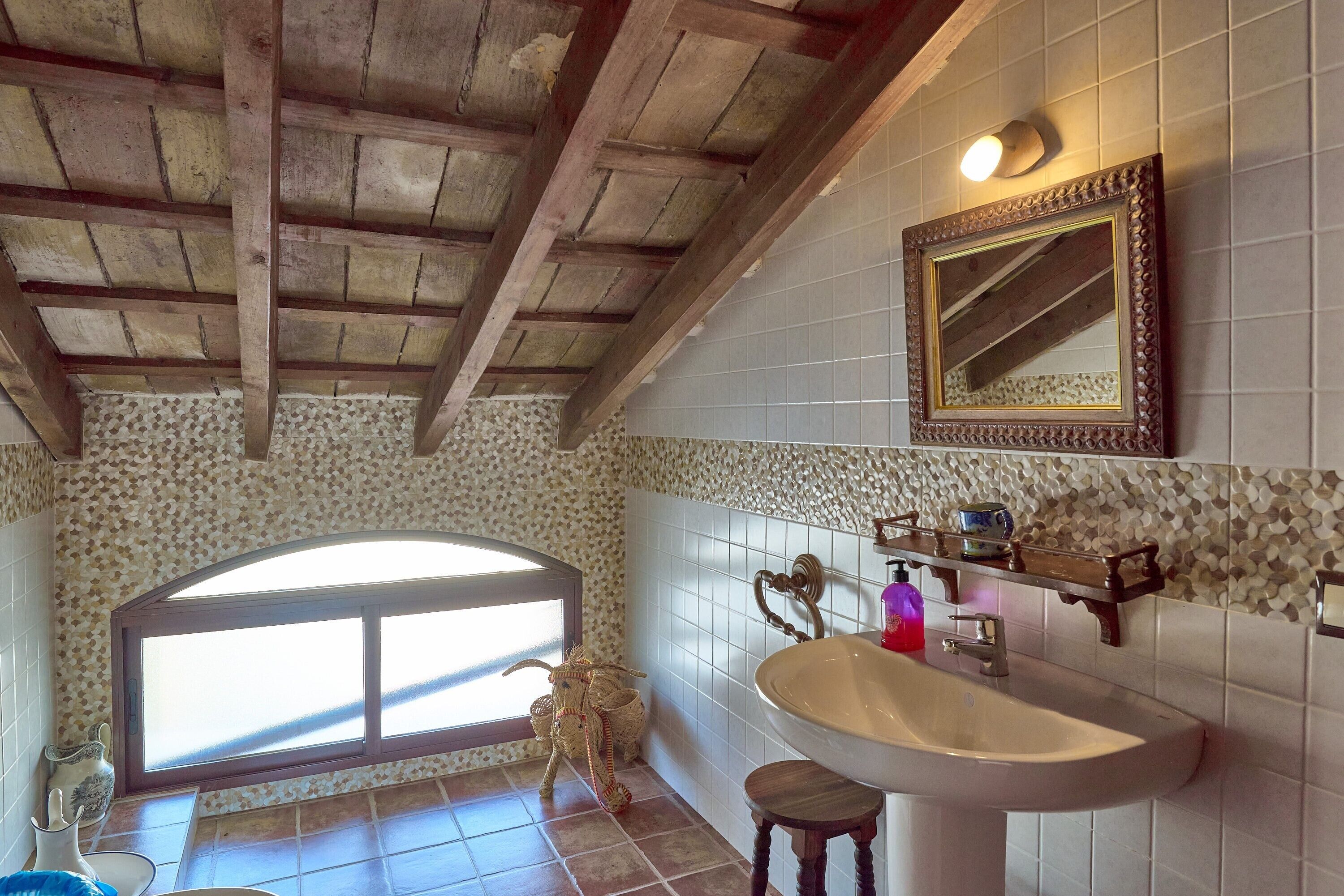 A bathroom with a wooden ceiling, a large window, a sink, a mirror, and a decorative vase.