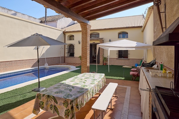 A patio with a pool, a table with a tablecloth, a grill, and a white canopy.