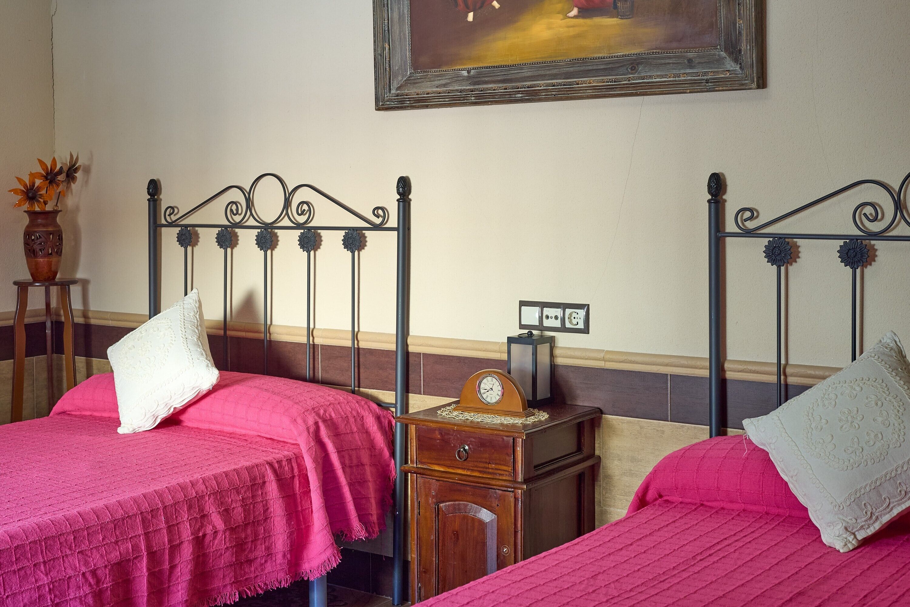 Two beds with metal frames, a wooden nightstand with a clock, and a vase with flowers on a side table.
