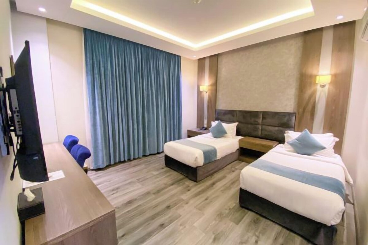A hotel room with two beds, a desk, a TV, and a large window with curtains.
