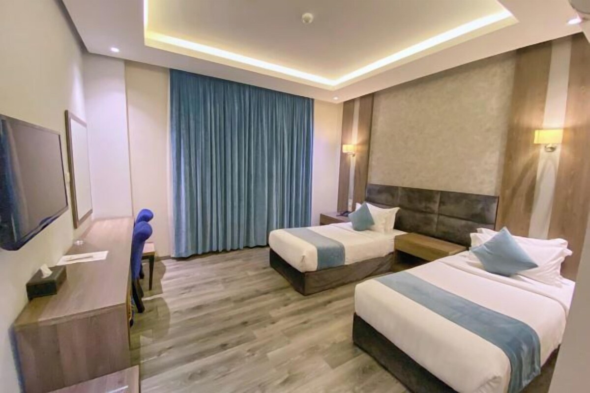 A hotel room with two beds, a desk, a TV, and a large window with curtains.