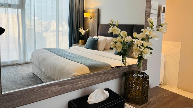 Ett modernt hotellrum med en stor säng, ett nattduksbord med en lampa, en vas med blommor, en spegel och utsikt över staden.
