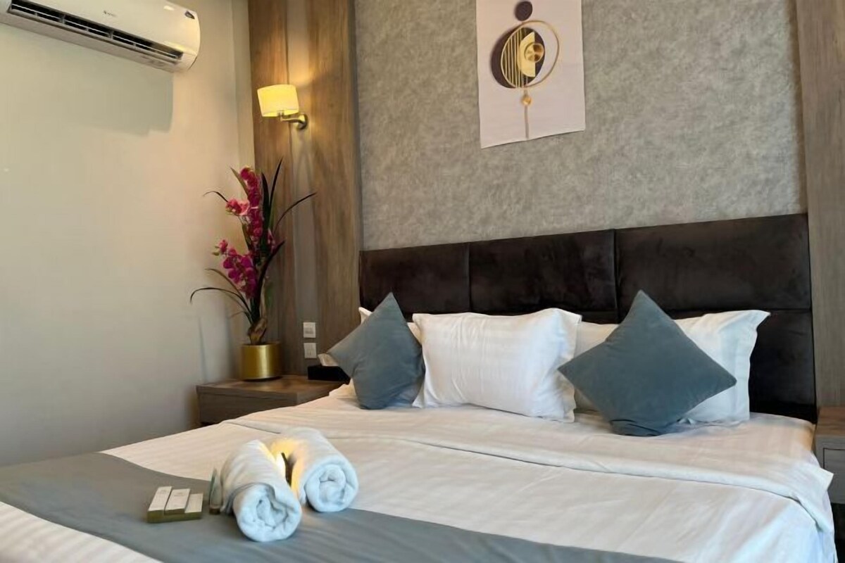 A hotel room with a bed, pillows, a nightstand, a lamp, and a wall-mounted air conditioner.