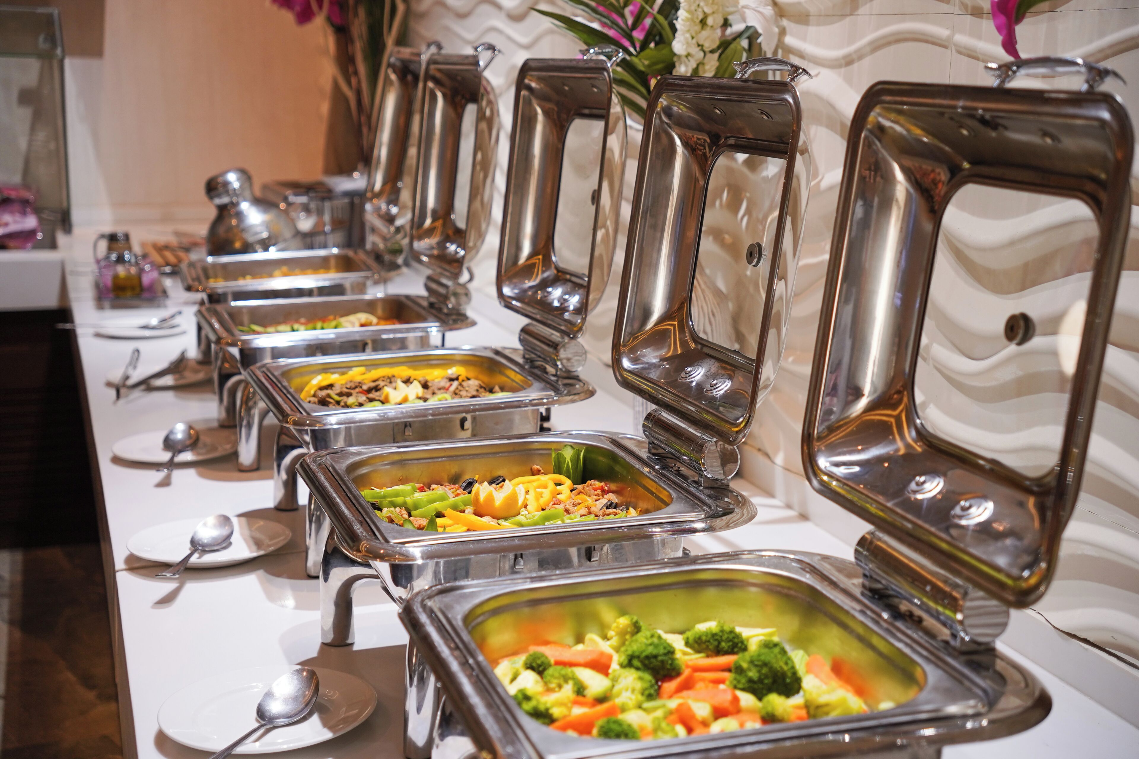 Una barra de buffet con diversas opciones de comida, incluyendo verduras y carnes, en recipientes de acero inoxidable.