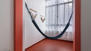 Eine Hängematte in einem Raum mit weißen Wänden und rotem Boden, nahe einem Fenster mit durchsichtigen Vorhängen.
